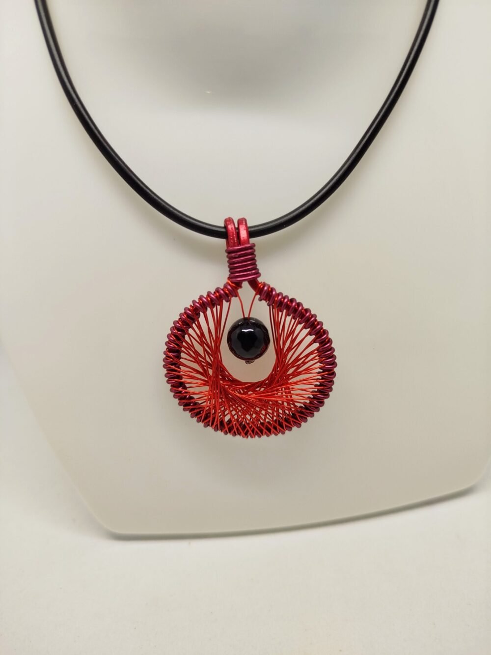 Le pendentif de 4 cm créé en fil aluminium rouge et tissé à l'intérieur. Une belle pierre Onyx à facettes est fixé au centre.Le collier de 45 cm de longueur est en caoutchouc avec fermoir mousqueton.
