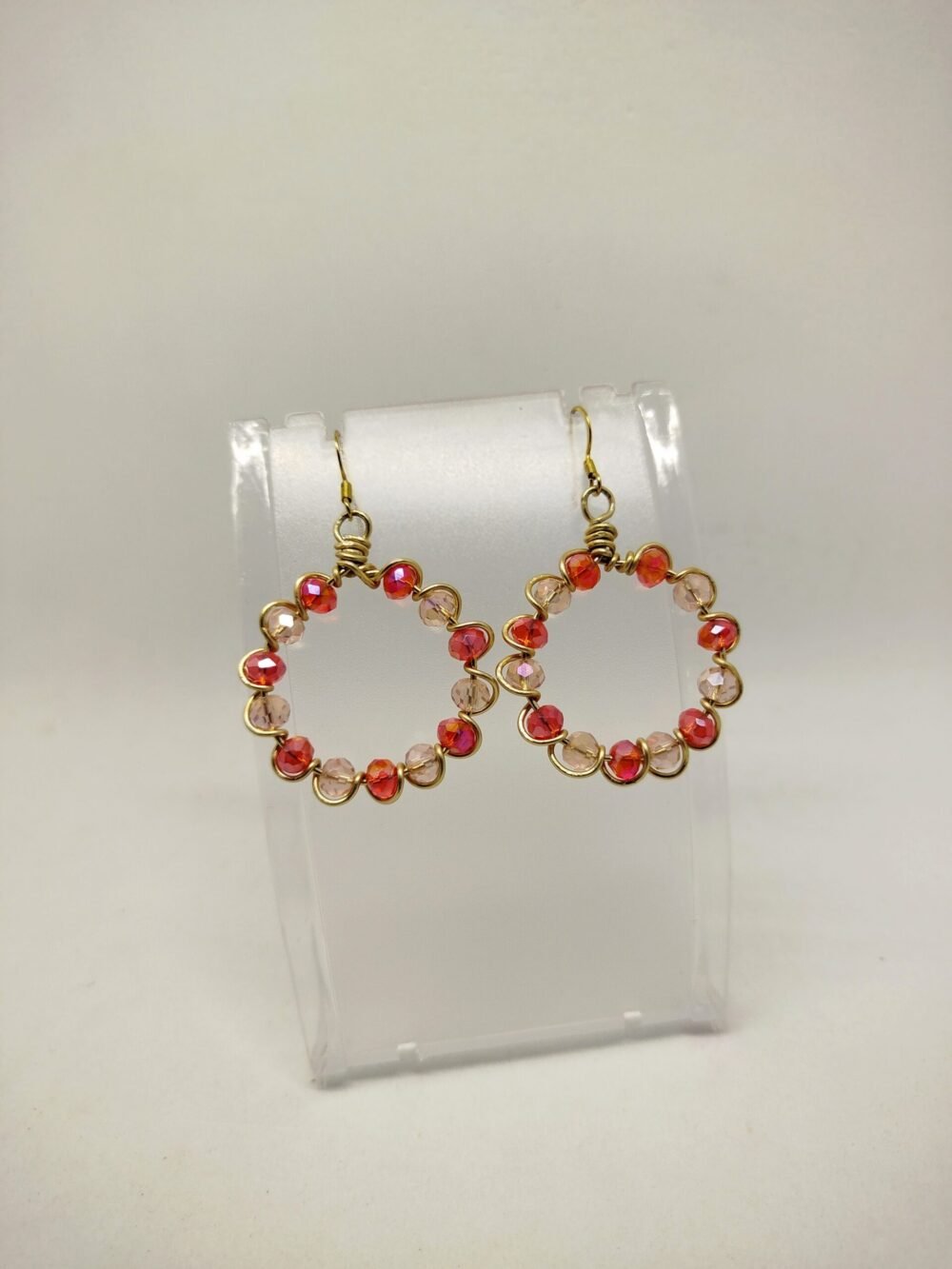 créoles façonnées avec des perles en verre à facettes rouge, entourées avec du fil aluminium doré. Leur diamètre est de 3 cm. Les crochets sont de couleur dorée et en acier chirurgical et hypoallergénique