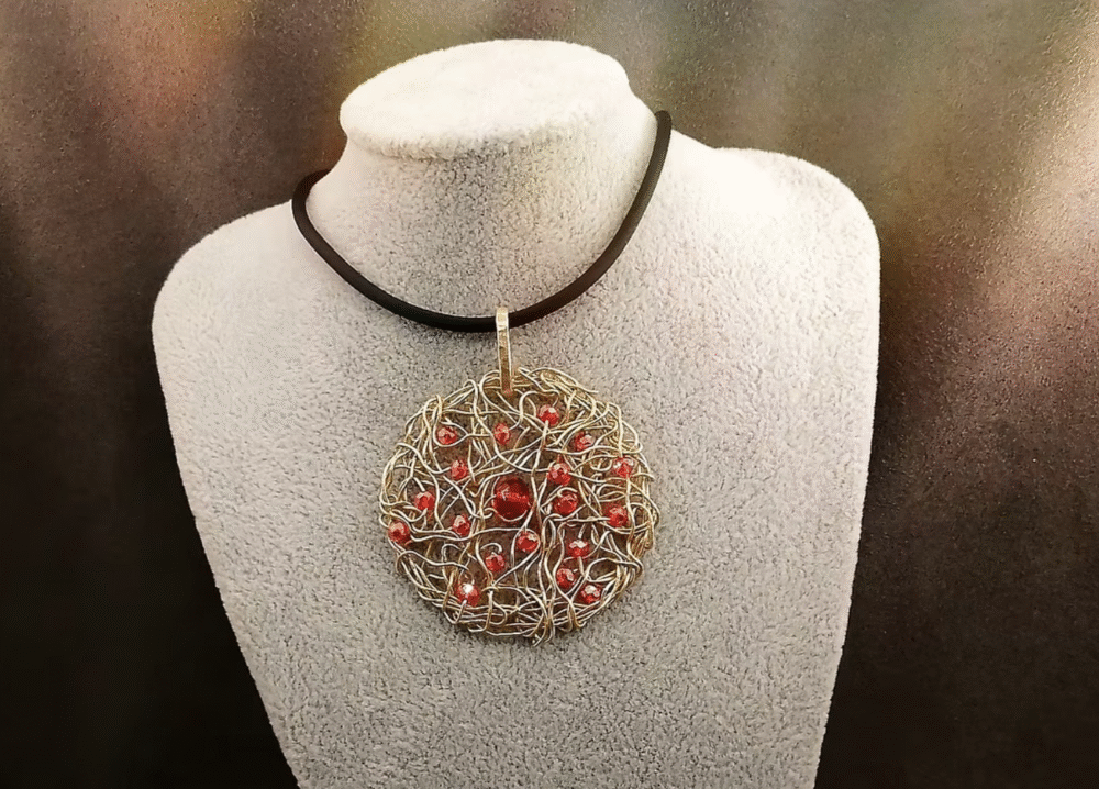 Le pendentif de 5cm confectionné à la main avec du fil aluminium doré et des petites perles à facettes rouges fixé sur un collier en caoutchouc noir d'une longueur de 50 cm avec fermoir mousqueton couleur doré.