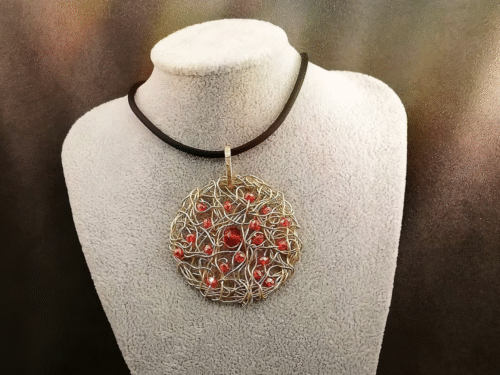 Le pendentif de 5cm confectionné à la main avec du fil aluminium doré et des petites perles à facettes rouges fixé sur un collier en caoutchouc noir d'une longueur de 50 cm avec fermoir mousqueton couleur doré.