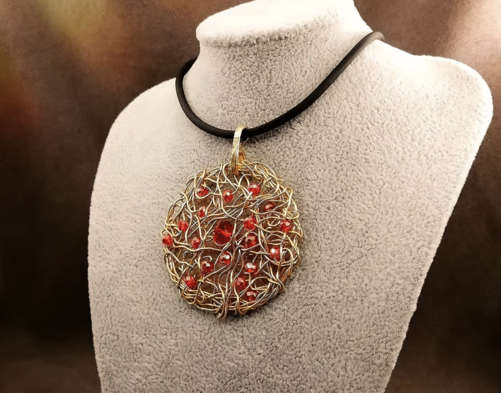 Le pendentif de 5cm confectionné à la main avec du fil aluminium doré et des petites perles à facettes rouges fixé sur un collier en caoutchouc noir d'une longueur de 50 cm avec fermoir mousqueton couleur doré.