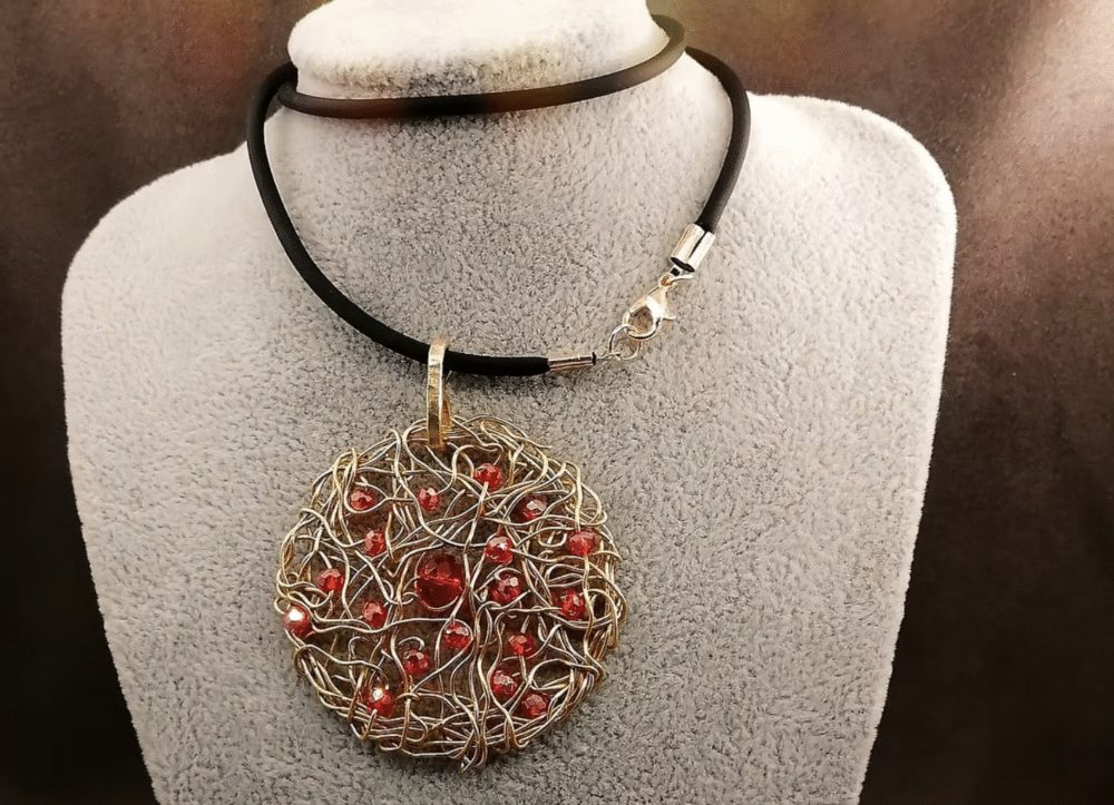 Le pendentif de 5cm confectionné à la main avec du fil aluminium doré et des petites perles à facettes rouges fixé sur un collier en caoutchouc noir d'une longueur de 50 cm avec fermoir mousqueton couleur doré.